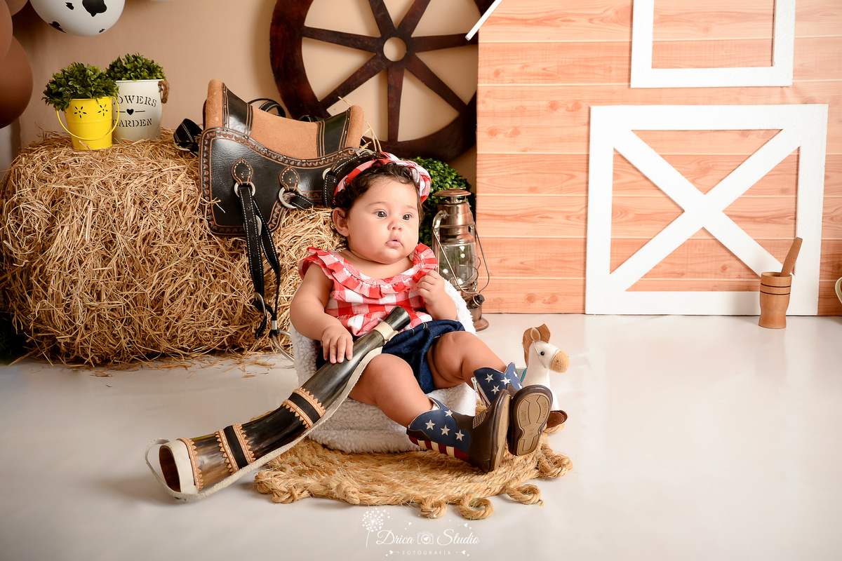 drica studio - ensaio fotografico infantil - fazendinha - xinguara - bebê senta em poltrona branca, vestindo blusa xadrez vermelha com branco e short jeans - berrante - rodão de carroça - feno - seleiro - balde amarelo com arbusto - pilão de madeira -sela