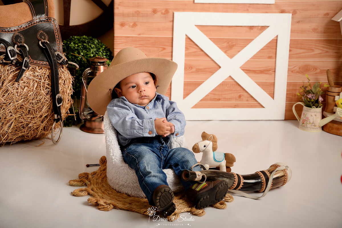 drica studio - ensaio fotografico infantil - fazendinha - xinguara -  bebê sentado em poltrona, vestindo camisa azul e calça jeana, bota morrom e chapéu - feno - sela - seleiro - berrante - cavalinho de pelucia - rodão de carroça - tapete rustico de corda