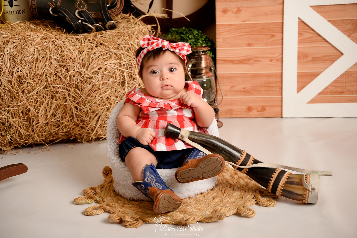 drica studio - ensaio fotografico infantil - fazendinha - xinguara - bebê sentada em poltrona, vestindo blusa xadrez vermelha com branco, segurando berrante - feno - sela - rodão de carroça - tapete rustico de corda - seleiro - lampião - bota marrom 