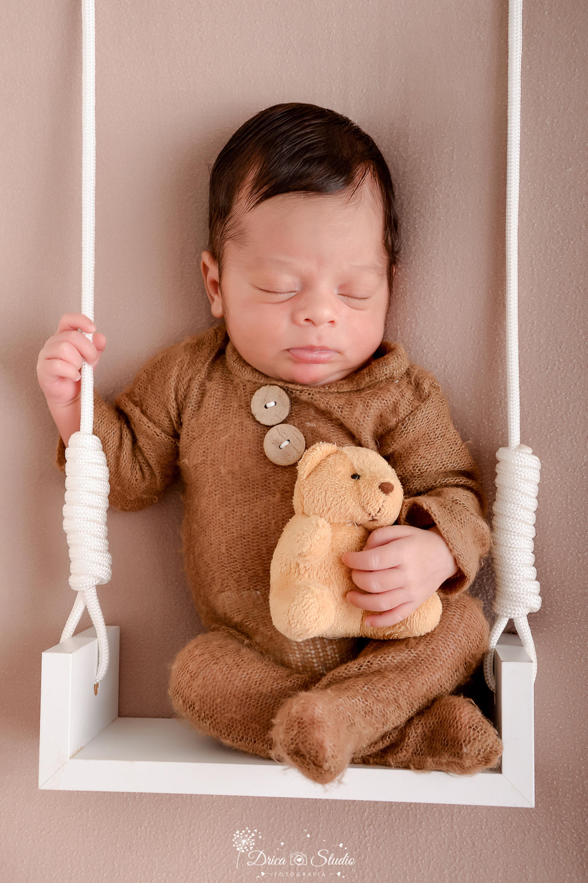 drica studio - ensaio fotográfico infantil - newborn - xinguara - bebê recém-nascido vestindo macacão marrom, sentado em balanço branco preso com cordas brancas e grossas, segurando um urinho de pelucia amarelo - fundo neutro