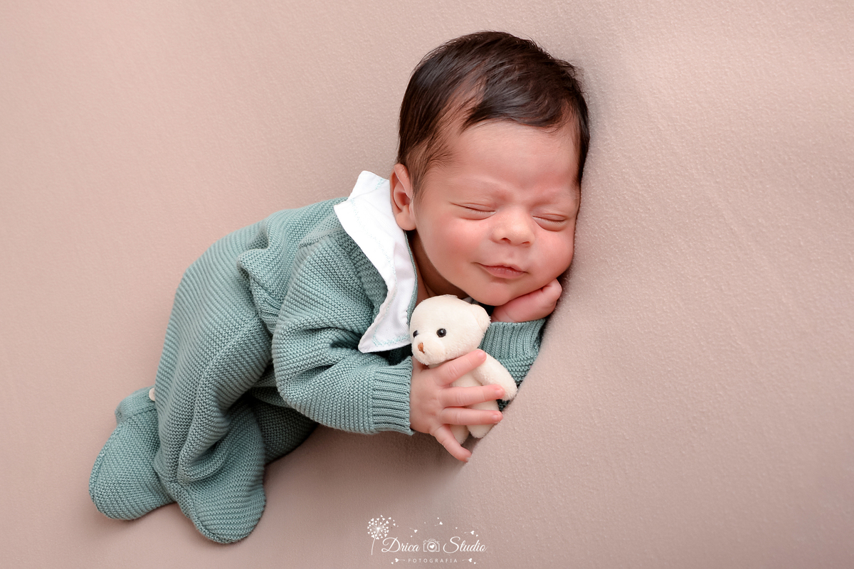 drica studio - ensaio fotográfico infantil - newborn - xinguara - bebê recém-nascido vestindo macacão de tricô verde pastel, deitado em superficie macia cor neutral, deitado de lado, segurando ursinho de pelucia branco.