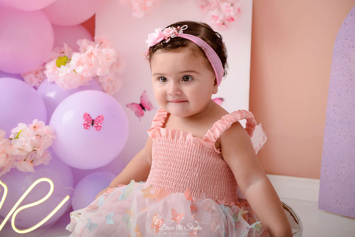 drica studio - ensaio fotografico infantil- smash the cake - jardim de borboletas - menina sentada em banquinho, usando vestido rosa com tule rosa claro com detalhes de borboleta, tiara de flores rosa - borboletas rosa - balões lilás e rosa - one de led 