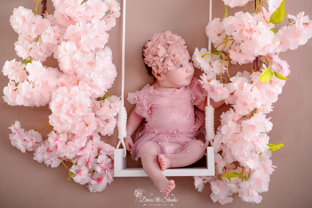 drica studio - ensaio fotografico infantil - newborn - xinguara - recém nascido sentada em balanço branco, com flores rosa em ambos os lado, vestindo bory rosa de renda e tule e tiara com flores de tule - fundo neutro