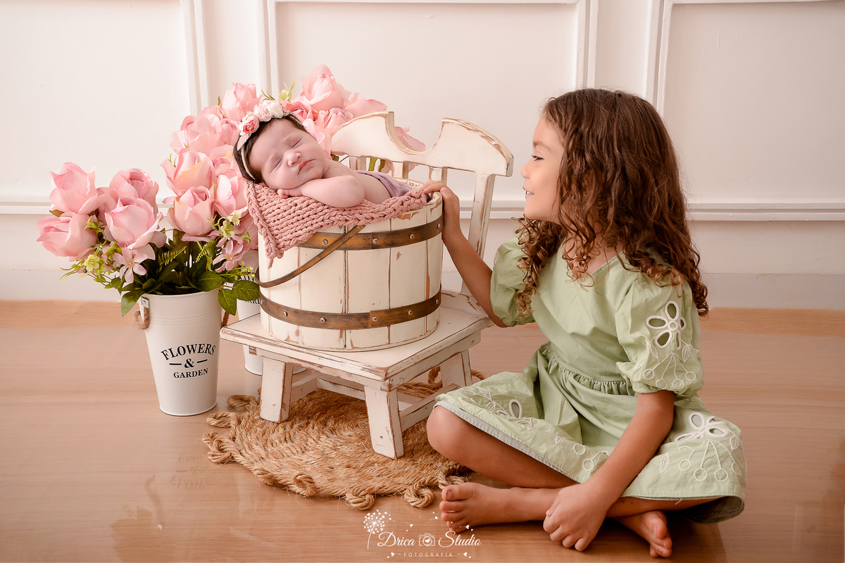 drica studio - ensaio fotografico infantil - newborn - xinguara - bebê dormindo dentro de balde branco rustico, sobre cadeira branca desgastada, ao lado da irmã mais velha que usa vestido vesde claro - vaso com flores rosa, tapete de corda rustico 
