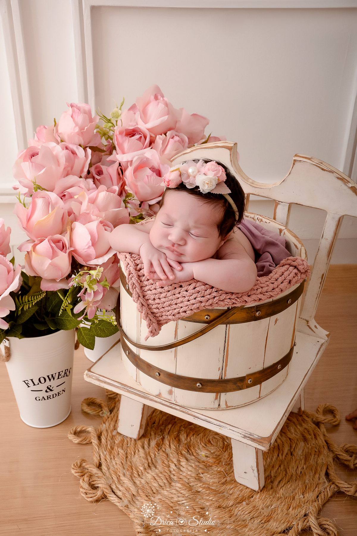 drica studio - ensaio fotografico infantil - newborn - xinguara - recém nascido dormindo dentro de balde de madeira rustico, acochoado com manta de tricô rosa, sobre cadeira de madeira branca desgastada, usando tiara de flores rosa na cabeça 