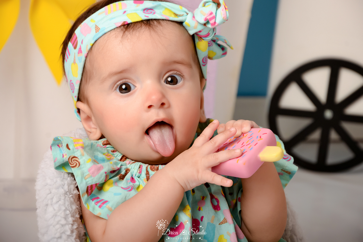 drica studio - ensaio fotográfico infantil - xinguara - acompanhamento mensal - mundo doce - bebê sentada em poltrona branca, vestindo bory e tiara com estampa de doces e fundo verde claro - carrinho de sorvete - fundo azul claro - pirulito e laço amarelo