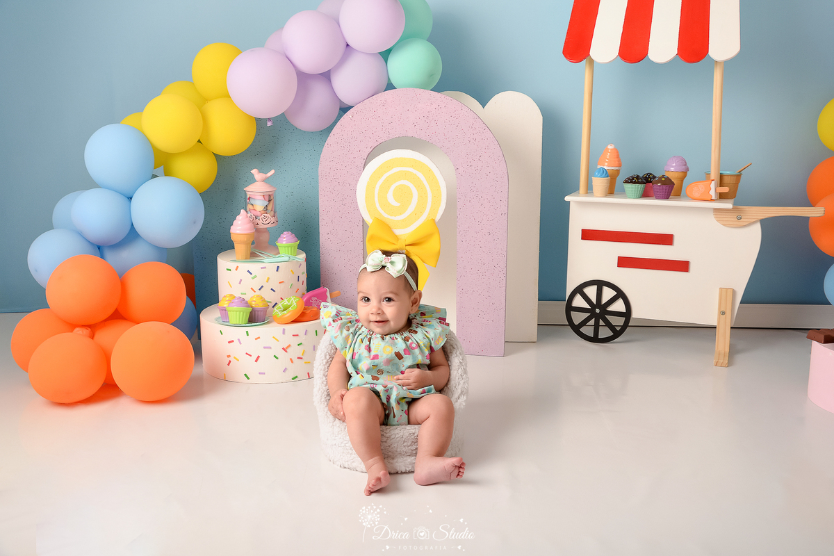 drica studio - ensaio fotográfico infantil - xinguara - acompanhamento mensal - mundo doce - bebê sentada em poltrona branca - balões coloridos - carrinho de sorvete - pirulito fake - bolo fake - arco lilás - fundo azul claro - guloseimas de brinquedo