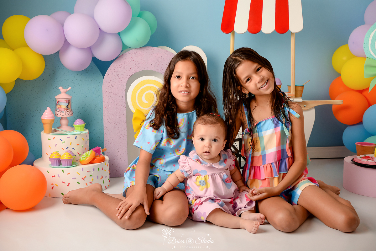 drica studio - ensaio fotográfico infantil - xinguara - acompanhamento mensal - mundo doce- três crianças sentada no chão, ao fundo  bolo fake, brinquedo em fotma de sorvete, bolinhos e doces - balões coloridos- carrinho de sorverte-pirulito