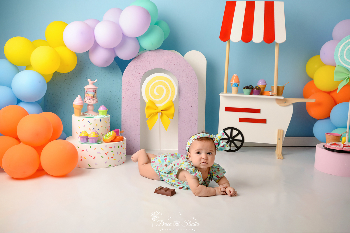 drica studio - ensaio fotográfico infantil - xinguara - acompanhamento mensal - mundo doce - bebê deitada no chão estindo bory e faixa de cabelo veste estampado - baloles coloridos - carrinho de sorvete branco com listras vermelhas - bolo branco fake 