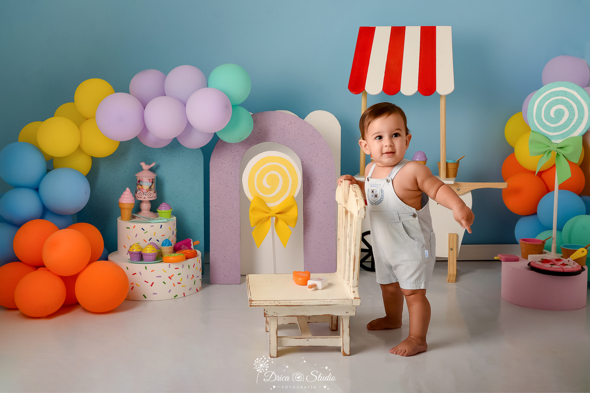 drica studio - ensaio fotográfico infantil - xinguara - acompanhamento mensal - mundo doce - bebê em pé apoiando em cadeira branca - pirulitos fake - sorverte, doces, tortas de brinquedo - balões coloridos - fundo azul claro - arco lilas