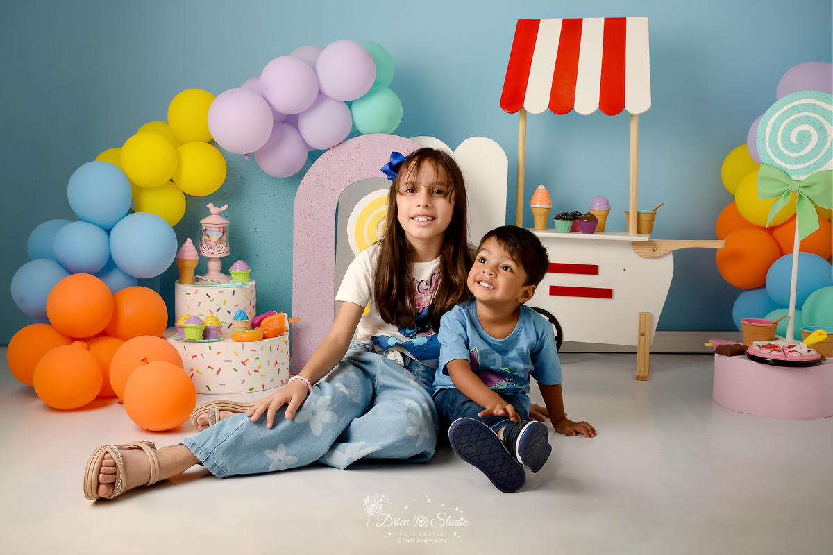 drica studio - ensaio fotográfico infantil - xinguara - acompanhamento mensal - mundo doce - casa de irmãos sentado do chão - ambos usam camisetas coloridas e calça jeans - carrinho de sorverte branco - balões coloridos - pirulito verde com laço verde