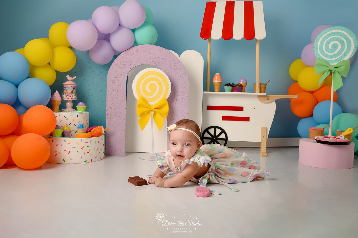 drica studio - ensaio fotográfico infantil - xinguara - acompanhamento mensal - bebê deitada no chão de bruços - carrinho de sorvete - bolo branco, pirulito amarelo e pirulito verde fake - sorverte, picoles, docinhos, rosquinhas de brinquedo - fundo azul 