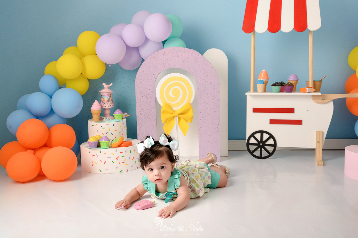 drica studio - ensaio fotográfico infantil - xinguara - acompanhamento mensal - mundo doce - bebê deitada no chão de bruco com lacinho no cabelo - carrinho de sorverte - pirulito fake - arco lilás - fundo azul claro - balões coloridos - bolo fake
