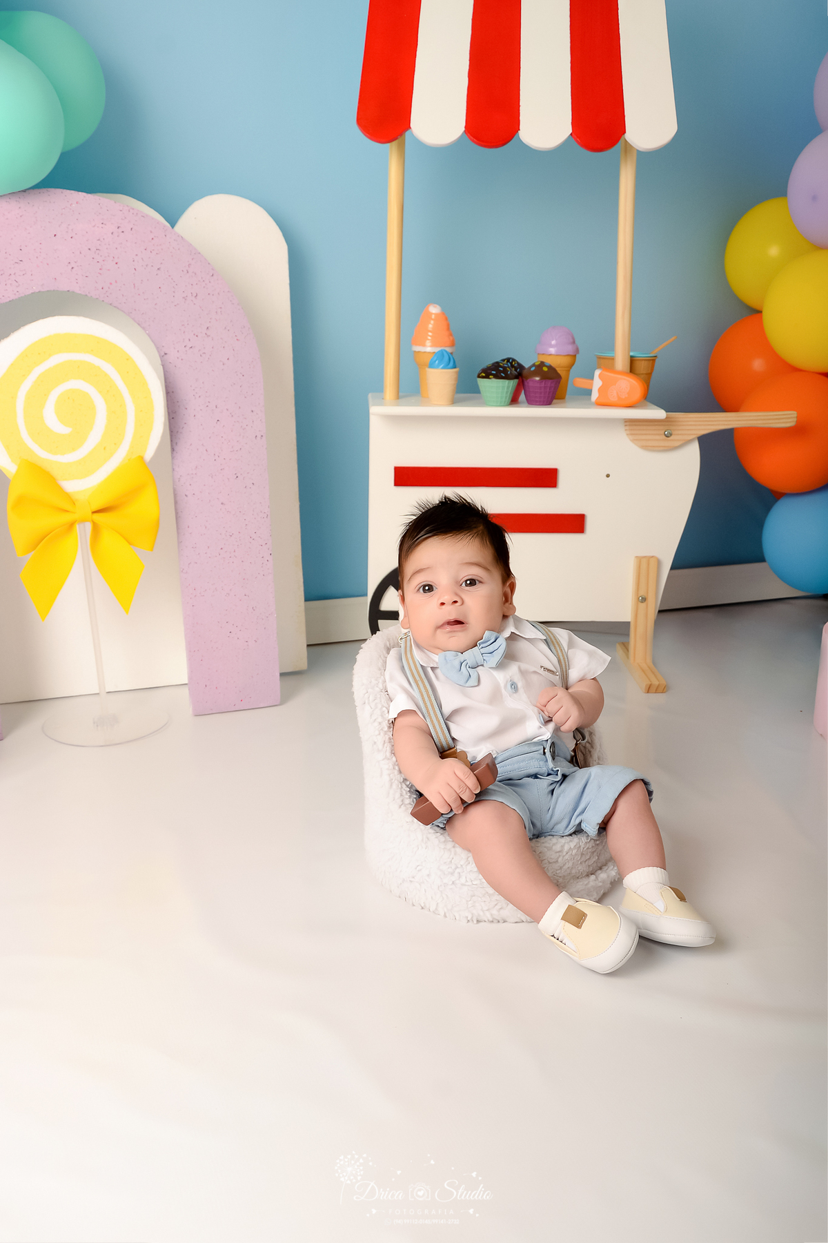 drica studio - ensaio fotográfico infantil - xinguara - acompanhamento mensal - mundo doce - bebê vestindo short e gravatinha azul claro e camisa branca, sentado em poltrona branca - pirulito amarelo - arco lilás - balão laranja - fundo azul claro - 