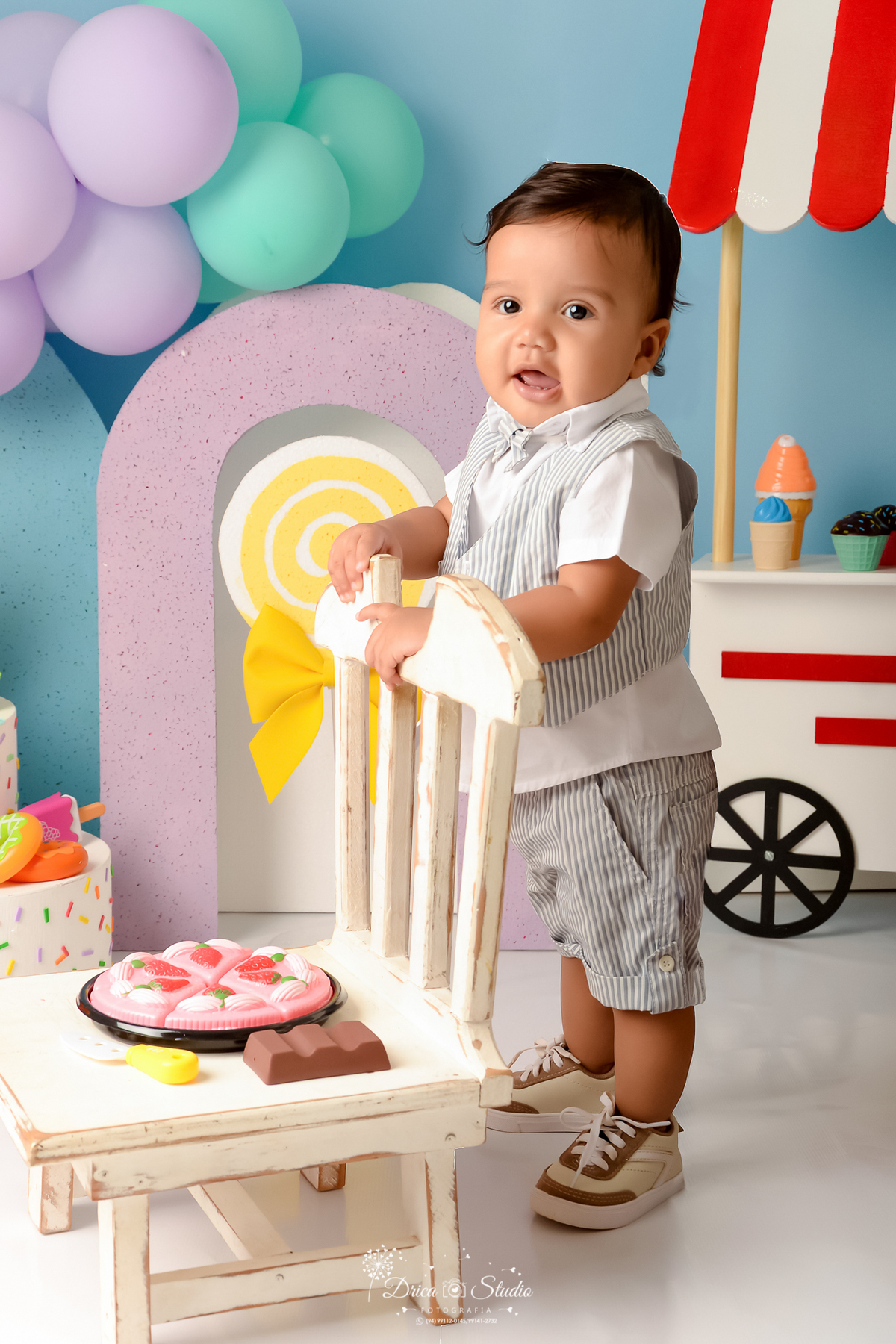 drica studio - ensaio fotográfico infantil - xinguara - acompanhamento mensal  - mundo doce - bebê em pé apoiado em cadeira branca, vestido short, colete e camisa - pirulito e laça amarelo - arco lilás - fundo azul - carrinho de sorvete branco brinquedo