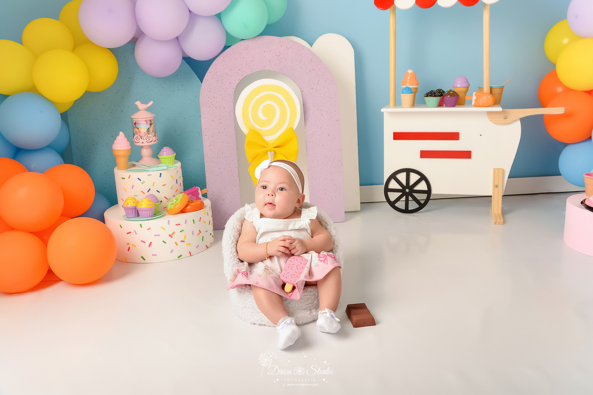 drica studio - ensaio fotográfico infantil - xinguara - acompanhamento mensal - mundo doce - bebê sentada em poltona, usando vestido branco com rosa - bolo branco com confetes coloridos - carrinho de sorvete branco com listras vermelhas - fundo azul claro