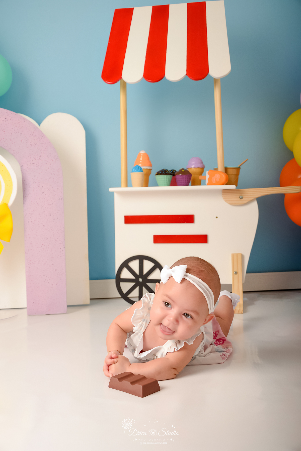 drica studio - ensaio fotográfico infantil - xinguara - acompanhamento mensal  - mundo doce - bebê deitada de bruços no chão - carrinho de sorvete branco com listras vermelhas - arco lilas - pirulito amarelo - fundo azul - balões coloridos -vestido branco
