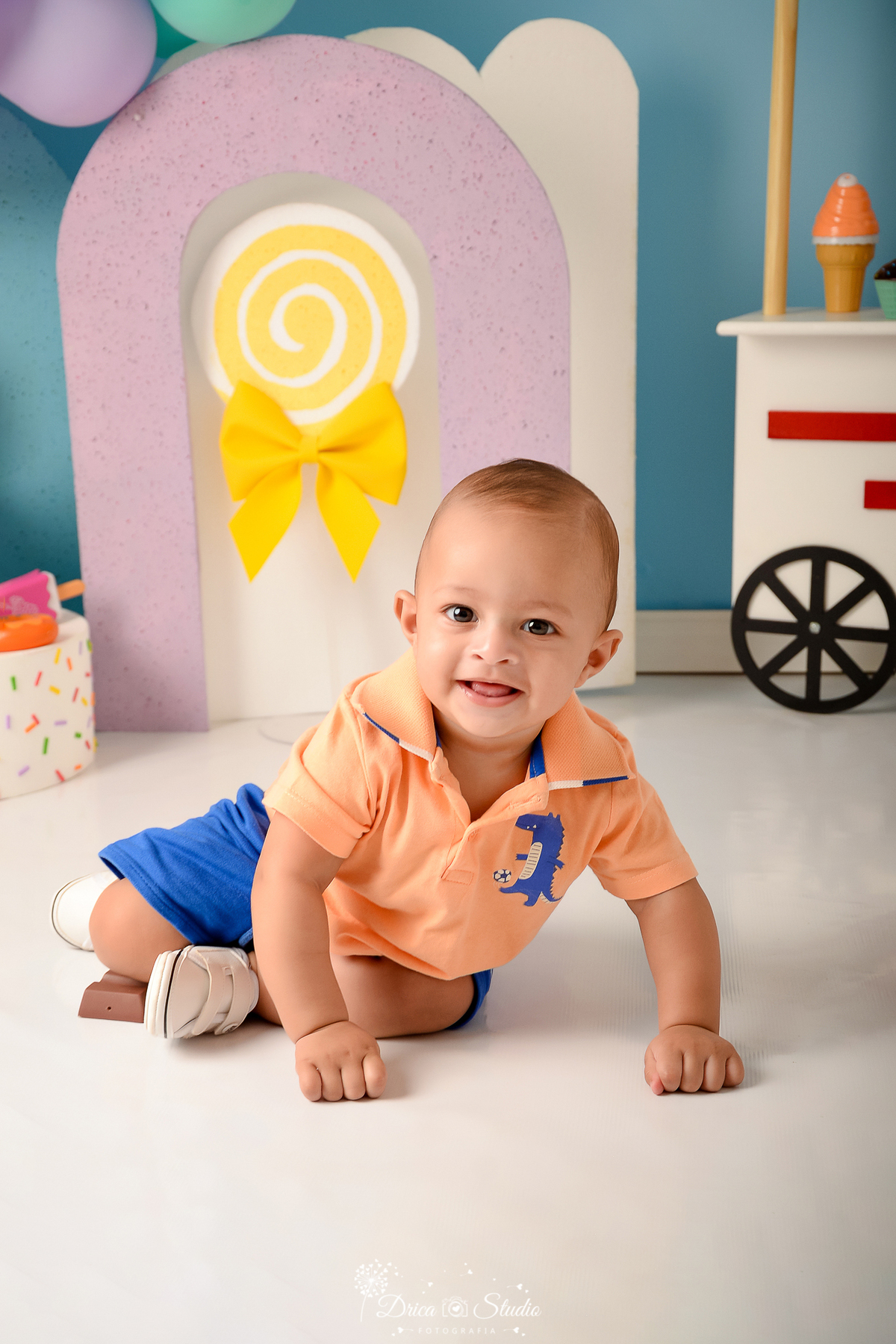 drica studio - ensaio fotográfico infantil - xinguara - acompanhamento mensal - bebê vestindo short azul escuro e camisa laranja com detalhe azul escuro - fundo azul claro - pirulito e laço amarelos - carrinho de sorvete - bolo com confetes coloridos