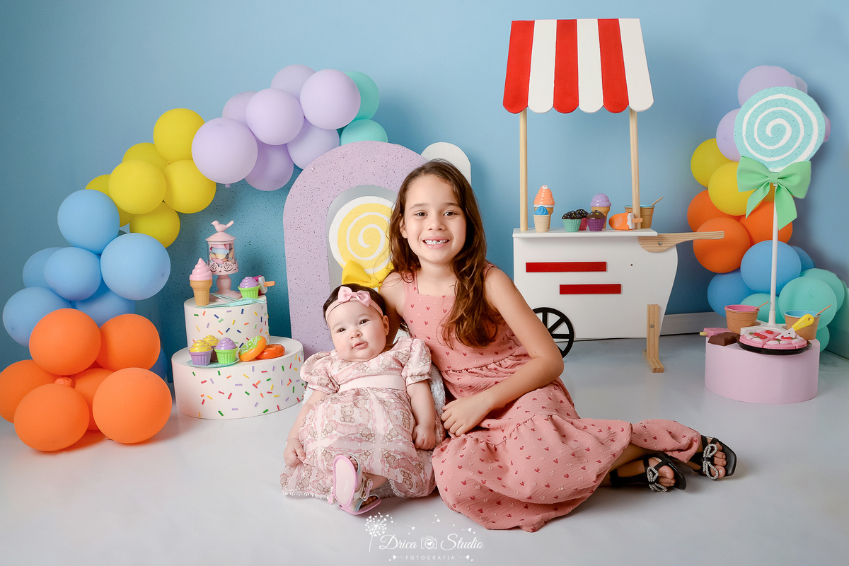 drica studio - ensaio fotográfico infantil - xinguara - acompanhamento mensal - bebê com vestido rosa de tule e renda e tiara com laço ao seu lado a irmã mais velha usando vestido coral - carrinho de sorvete - arco lilás - balões coloridos -pirulito verde