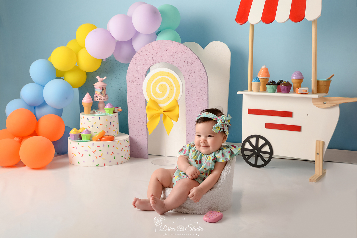 drica studio - ensaio fotográfico infantil - xinguara - acompanhamento mensal - bebê sentada em poltrona, vestindo bory e faixa de cabelo verde com estampas - carrinho de sorvete - balões coloridos - pirulito amarelo - bolo branco com confete coloridos
