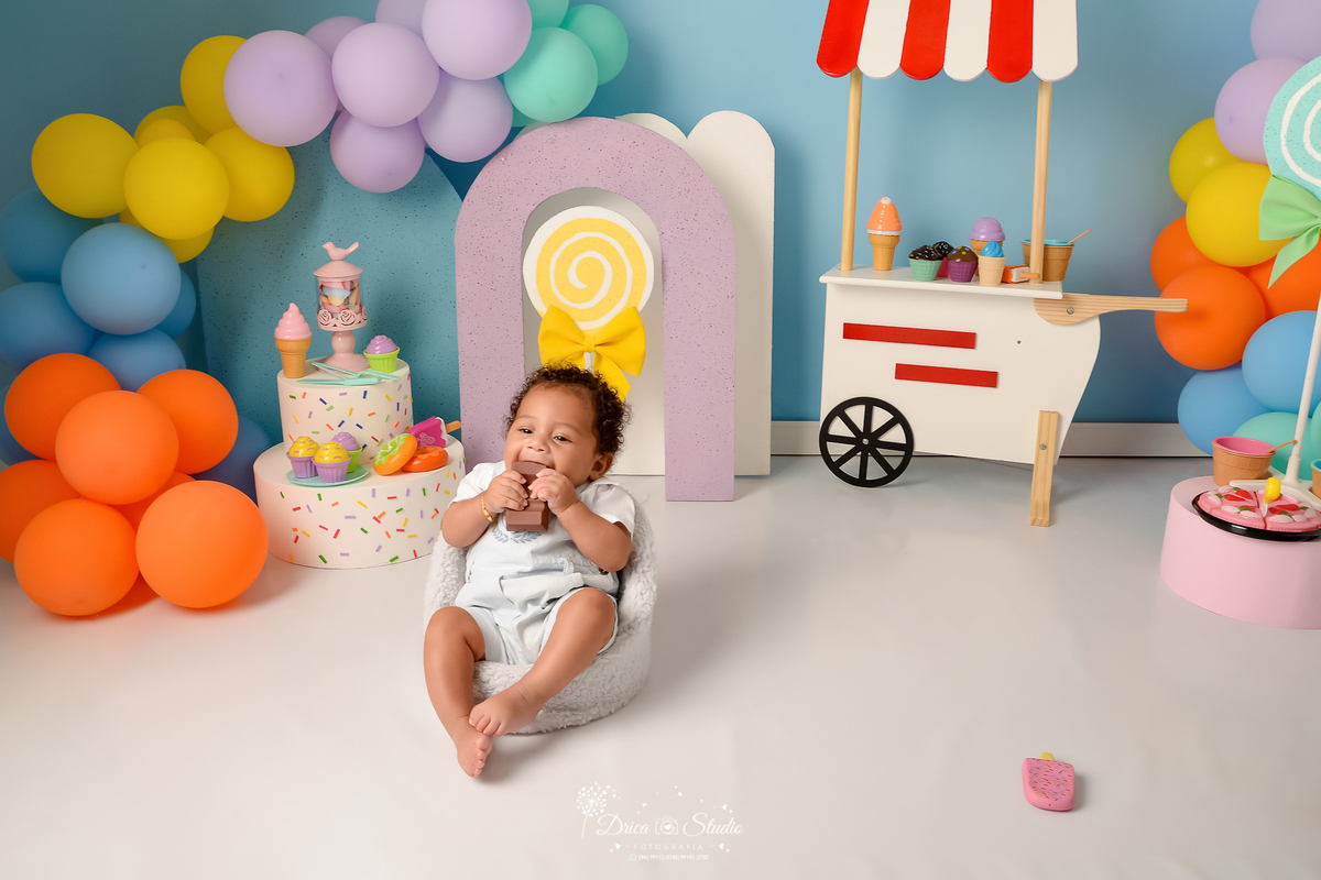 drica studio - ensaio fotográfico infantil - xinguara - acompanhamento mensal - mundo doce - bebê vestindo macaquinho azul claro com camisa branca, mordendo chocolate de brinquedo - carrinho de sorvete branco com listras vermelhas - balões coloridos 
