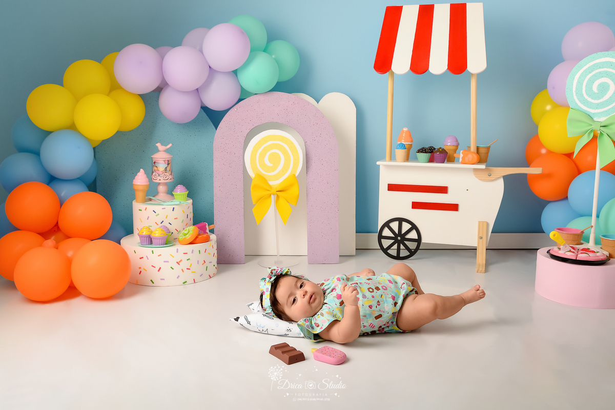drica studio - ensaio fotográfico infantil - xinguara - acompanhamento mensal - mundo doce - bebê deitada no chão com cabeça sobre travesseiro - balões coloridos - bolo branco com confetes coloridos - pirulitos amarelo e verde - carrinho de sorvete branco