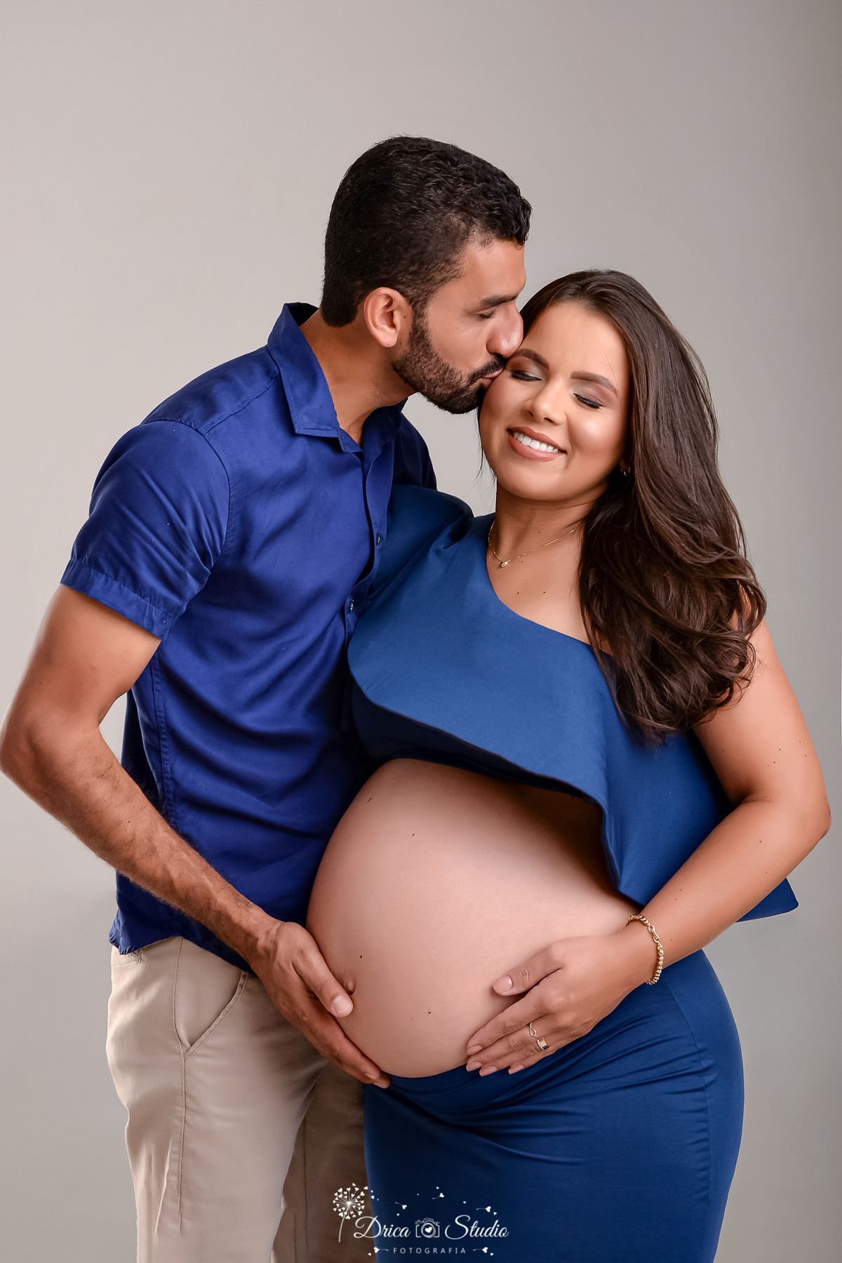 drica studio - ensaio fotográfico - gestante -xinguara- gestante usa conjunto azul escuro, blusa deixa a barriga a mostra, ao seu lado o pai beijando-a no rosto e com a mão sobre a barriga da esposa gestante - fundo neutro