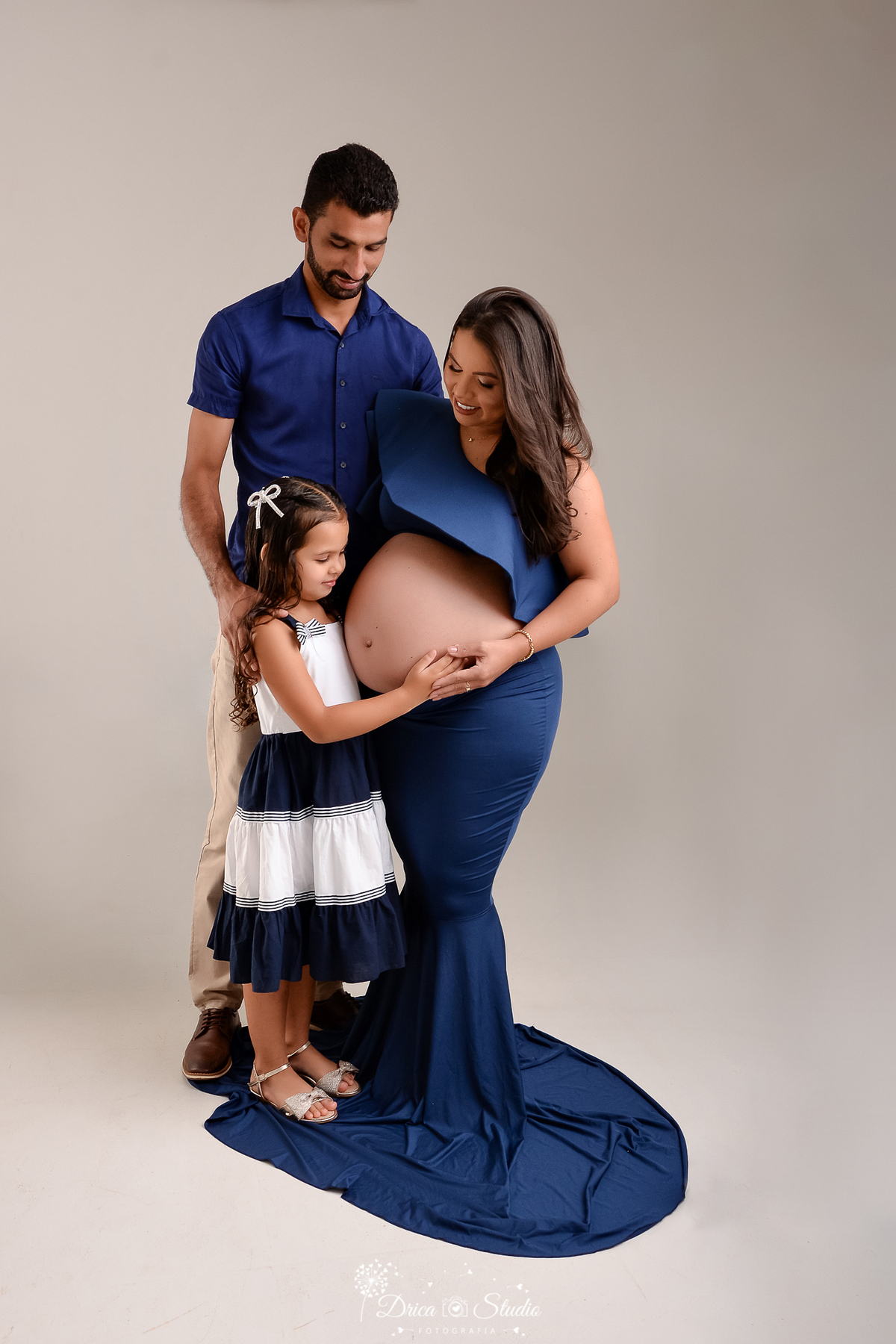 drica studio - ensaio fotográfico - gestante- A mãe, vestindo um conjunto azul que destaca a barriga de grávida,O pai, de camisa azul e filha usando um vestido azul e branco,toca com delicadeza a barriga da mãe - fundo neutro