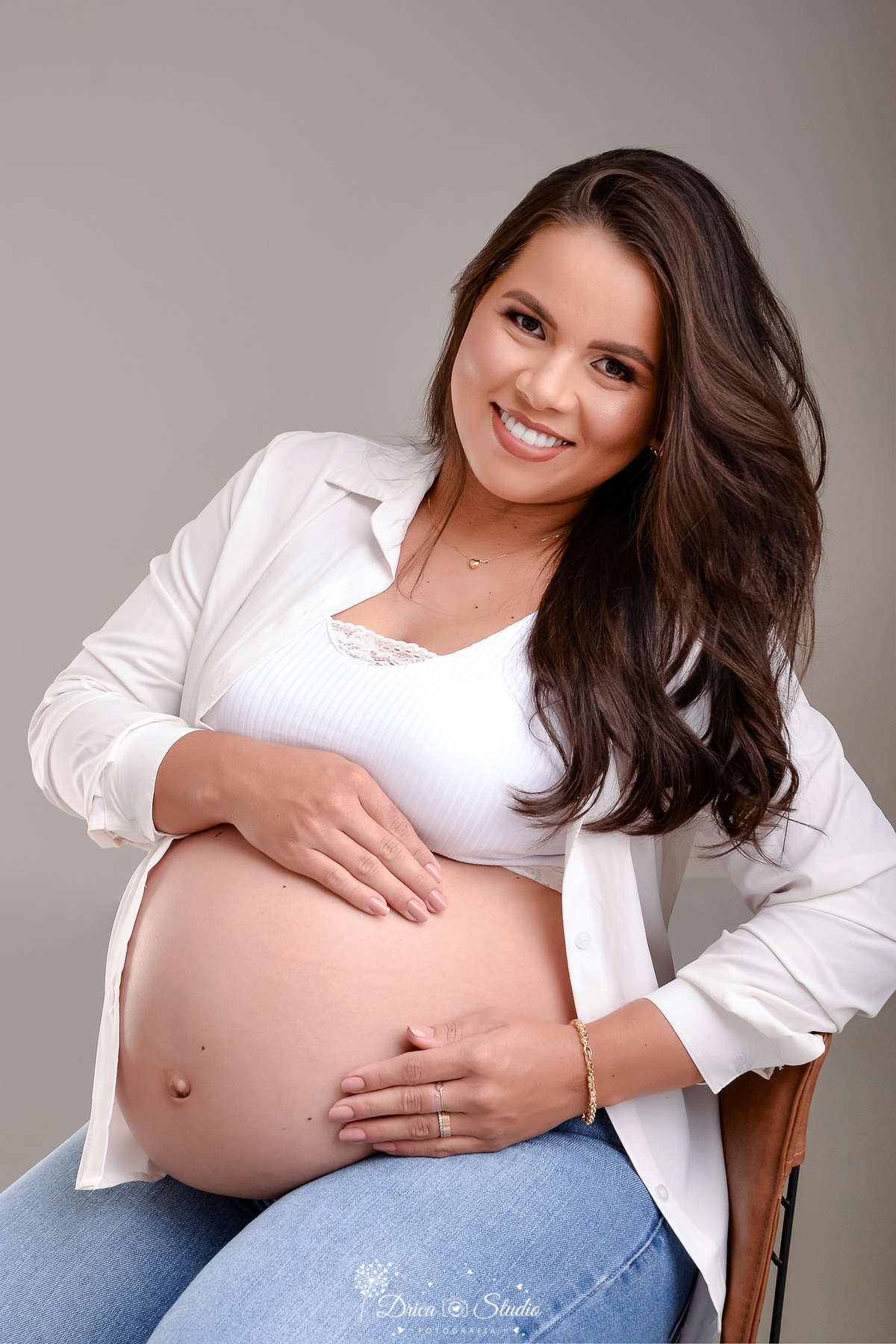 drica studio - ensaio fotográfico - gestante -xinguara- gestante sentada em cadeira de madeira com acento em couro marrom, veste calça jeans, camisa e cropped branco, e estar com braça apoiado sobre a barriga que está a amostra- fundo neutro