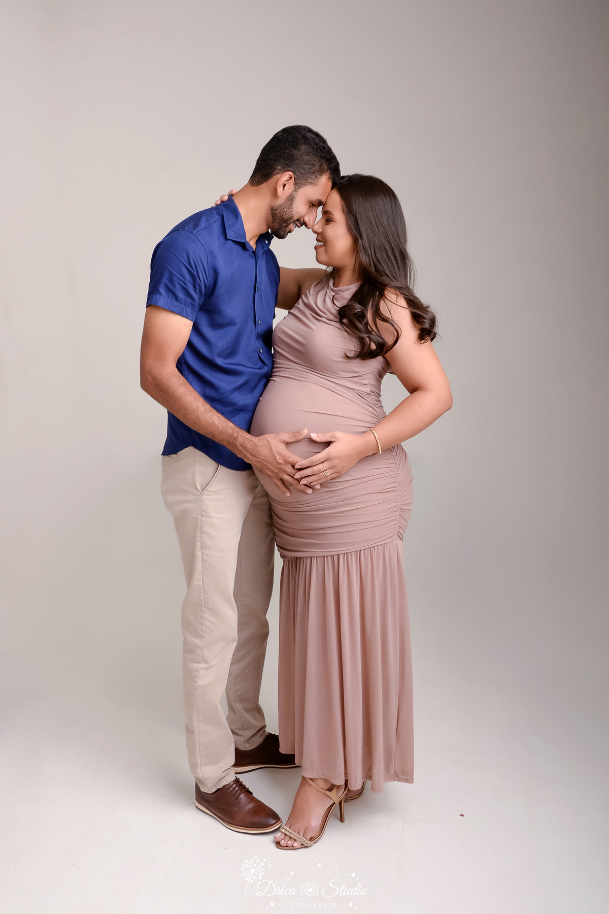 drica studio - ensaio fotográfico - gestante -xinguara- gestante-casal frente a frente, com os rostos encostados em um gesto de carinho e cumplicidade. O pai veste uma camisa azul e calça clara, enquanto a mãe usa um elegante vestido longo nude