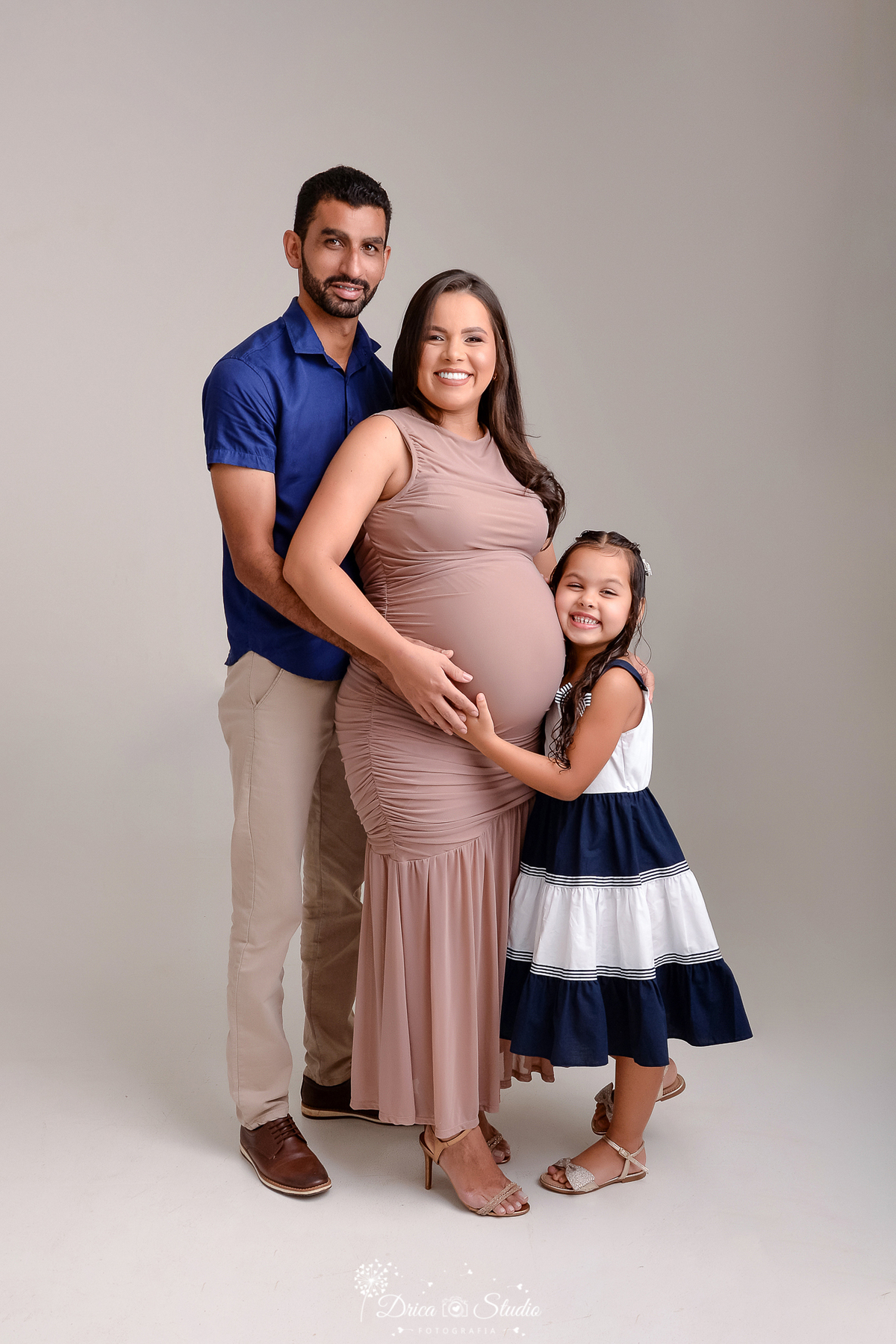 drica studio - ensaio fotográfico - gestante -xinguara- gestante usando vestindo nude, a direita a filha a abraça e encosta o rosta na barriga da mãe, a esquerda o pai usando camisa azul escuro e calça bege- fundo neutro
