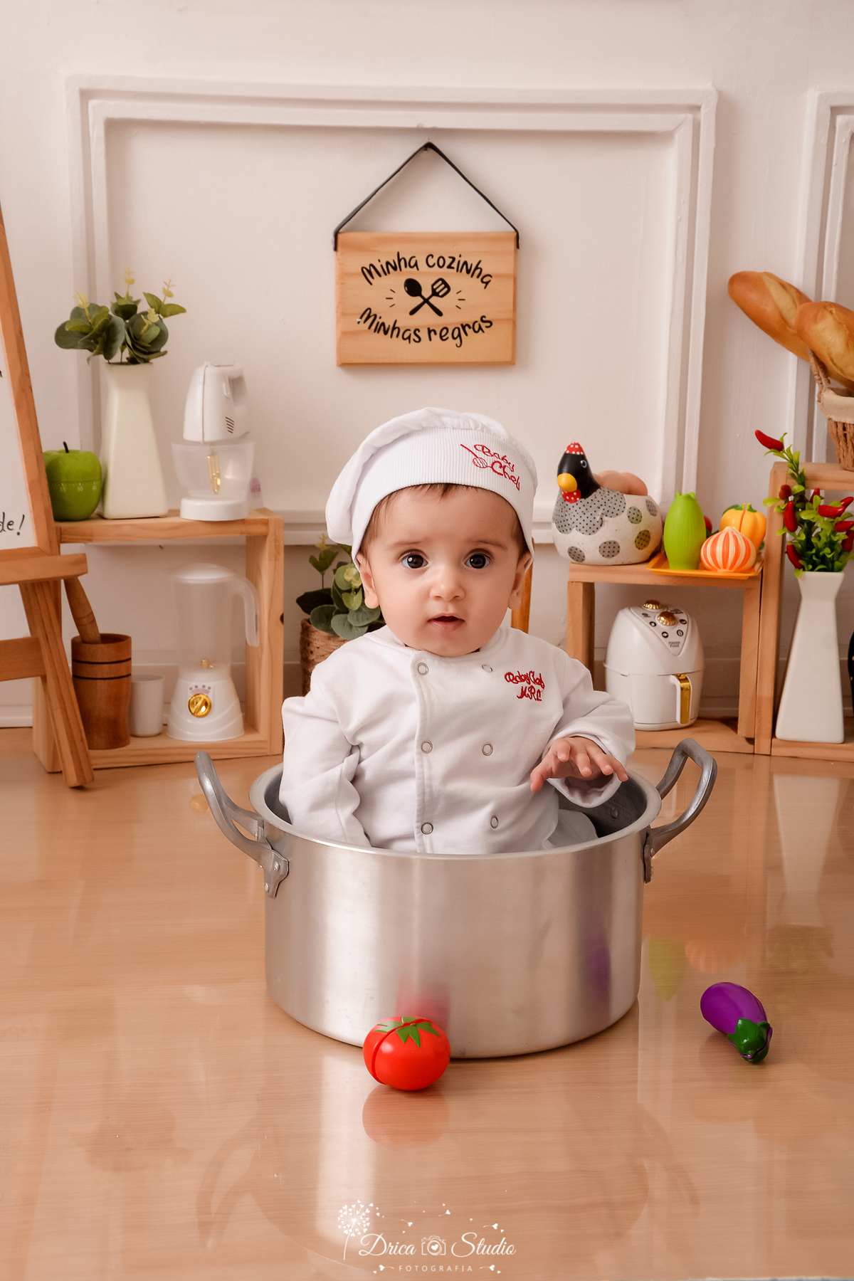 drica studio - ensaio fotográfico infantil - acompanhamento - bebê sentada dentro de panela, vestida com roupa de chef de cozinha - legumes e galinha com ovos decorativos - fundo branco com boiserie e piso amadeirado