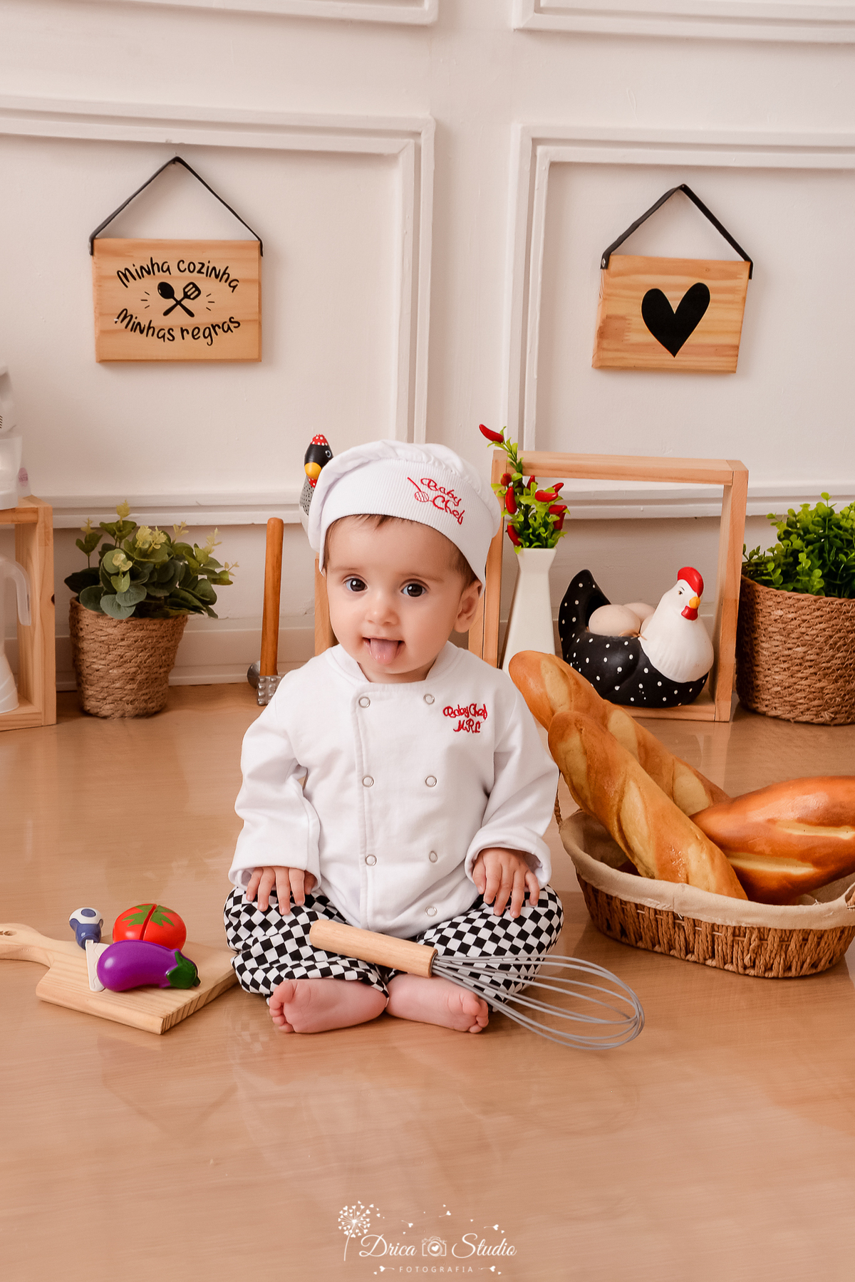drica studio - ensaio fotográfico infantil - acompanhamento - bebê sentada no chão, vestida de chef de cozinha, ao seu lado um cesto de pães - cesto com artustos - galinha decorativa - fundo branco com boiserie e piso amadeirado