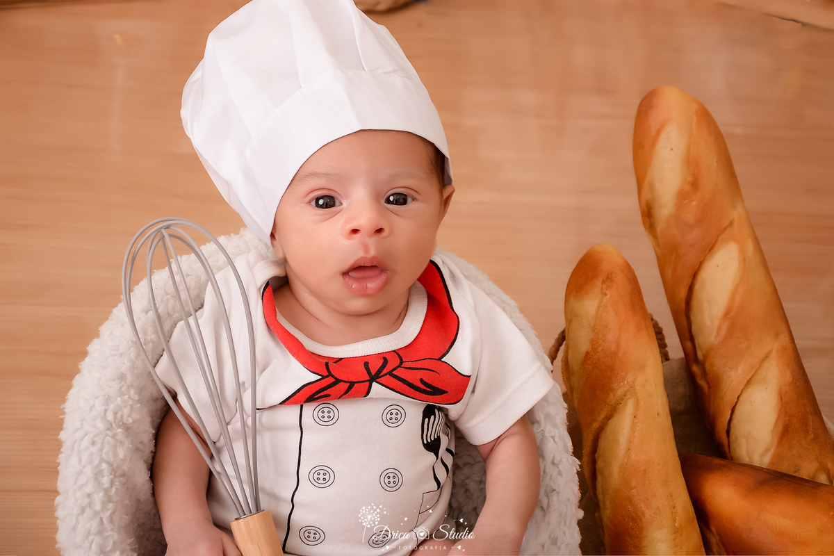 drica studio - ensaio fotográfico infantil - acompanhamento mensal - vestido de pequeno chef de cozinha, usando um chapéu de cozinheiro branco, segurando um batedor de massa- pães dentro de um cesto - piso amadeiradovestido de pequeno chef de cozinha, usa