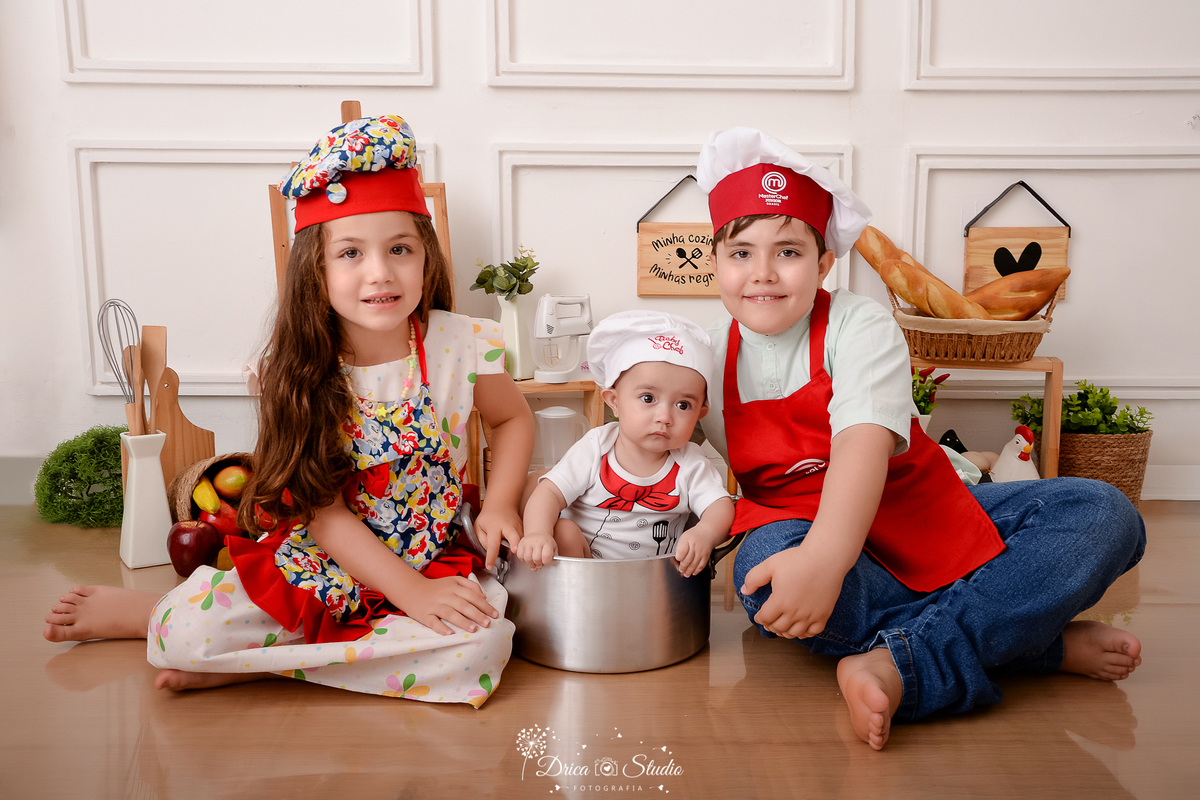 drica studio - ensaio fotográfico infantil - acompanhamento - bebê sentada dentro de panela, do lado direito o irmão e do esquerdo a irmã, ambos vestidos de chef de cozinha - utensilios de cozinha - fundo branco com boiserie e piso amadeirado