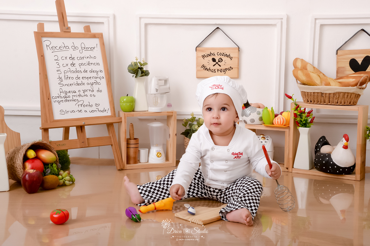 drica studio - ensaio fotográfico infantil - acompanhamento mensal- bebê sentado no chão vestindo roupa de chef de cozinha - frutas dentro de cesto de palha - utensilios de cozinha - galinha com ovos decorativo - quadros decorativos - fundo branco 