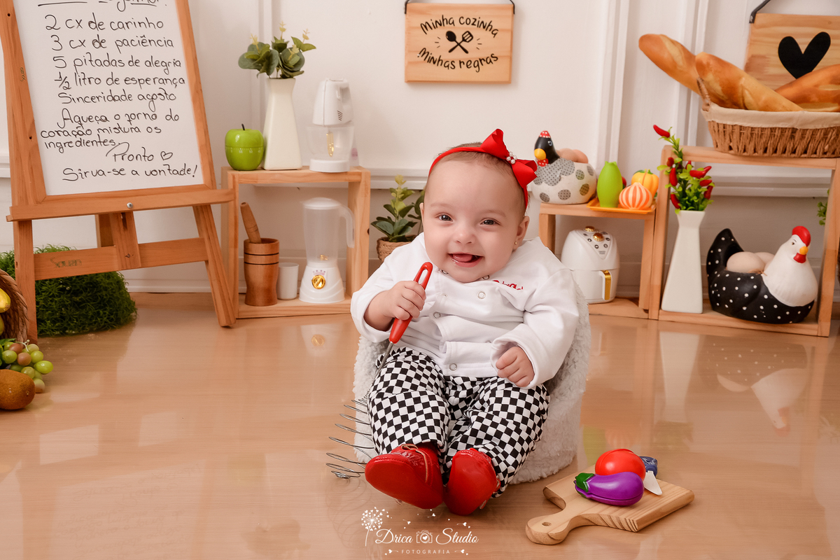 drica studio - ensaio fotográfico infantil - acompanhamento mensal- bebê sentando em poltrona branca vestindo roupa de chef de cozinha - tabúa de cortar- berinjela e tomate - utensilios de cozinha - vas branco com folhagem - pães no cesto de palha - 