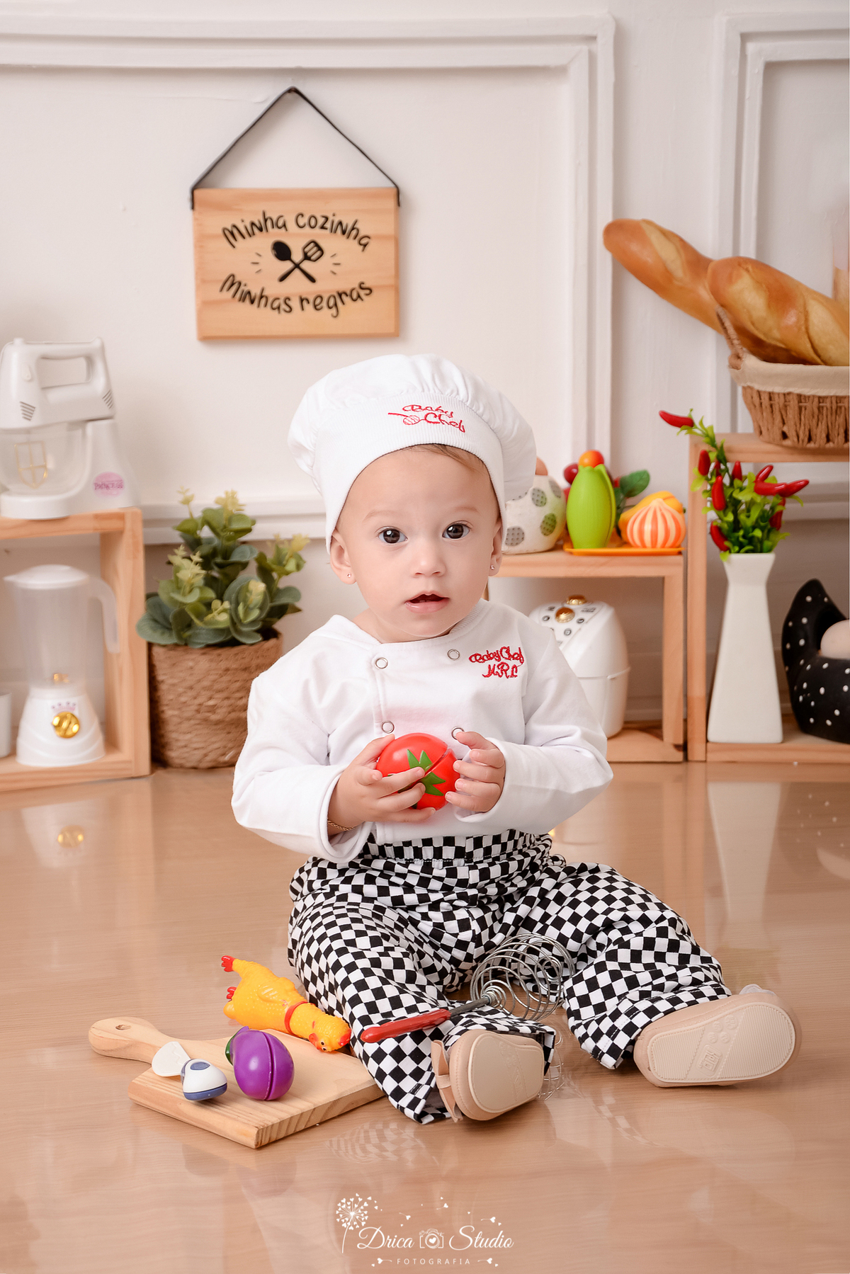 drica studio - ensaio fotográfico infantil - acompanhamento - bebê sentada no chão, vestida de chef de cozinha, segurando um tomate a sua frete - utesilios de cozinha - liquidificador - legumes e galinha com ovos decorativos - fundo branco 