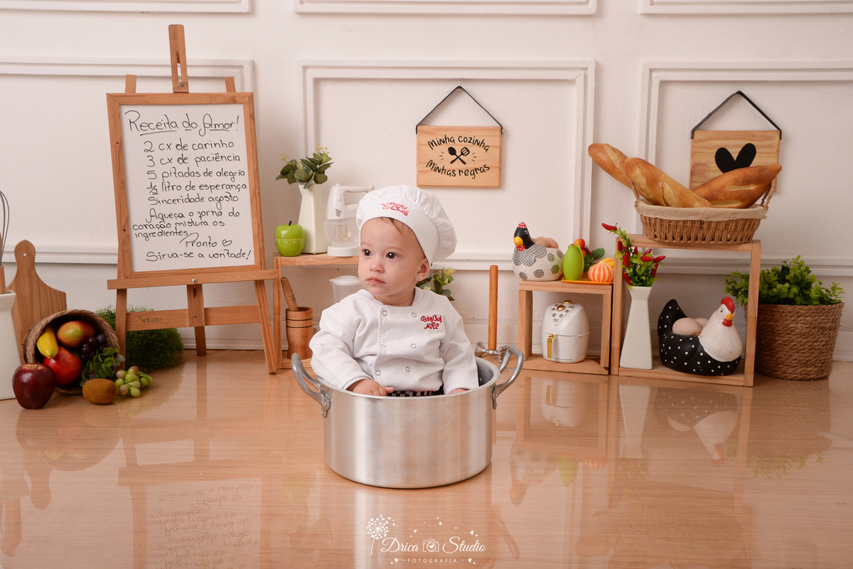 drica studio - ensaio fotográfico infantil - acompanhamento - bebê sentado dentro de panela - quadrinhos decorativos - placa com receita - cesto com frutas - galinha decorativa - utensilios de cozinha - fundo branco com boiserie e piso amadeirado