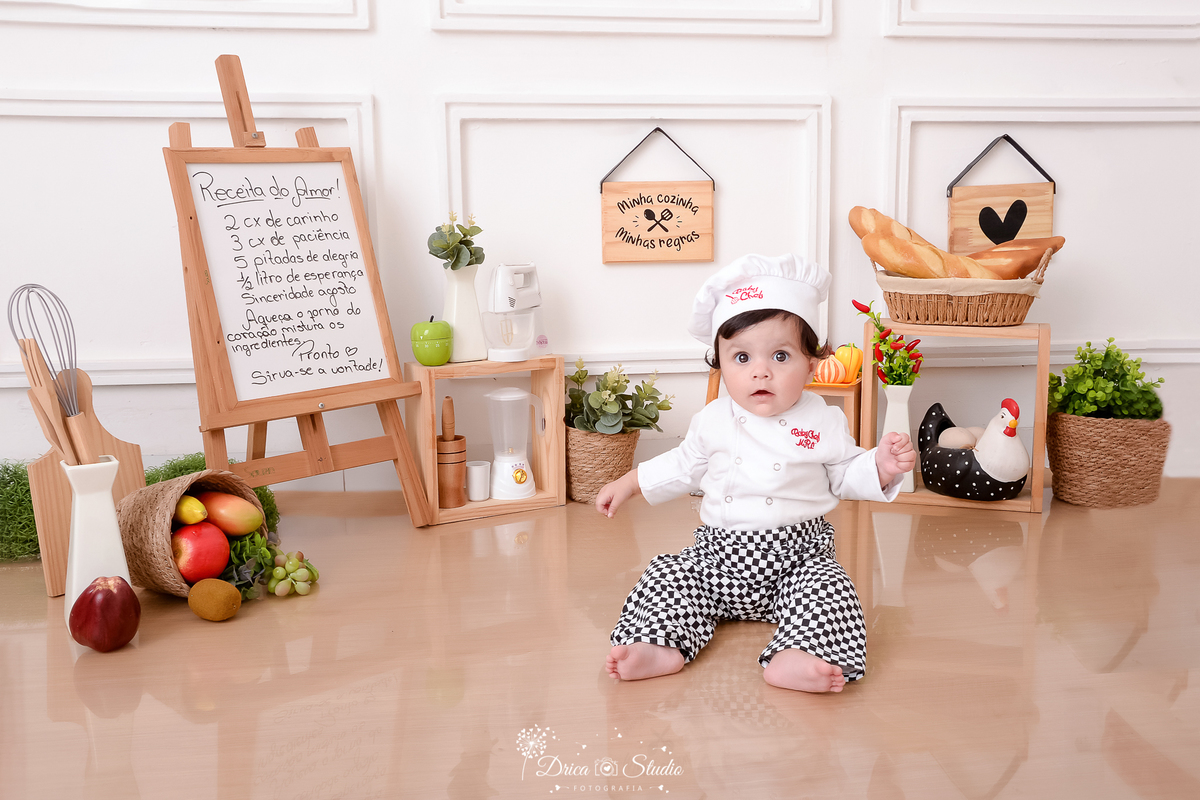 drica studio - ensaio fotográfico infantil - acompanhamento - bebê sentada no chão vestindo roupa de chef de cozinha - cesto com pães - cesto com frutas - placa com receita - cesto com arbusto - liquidificador - panela eletrica - galinha decorativa 