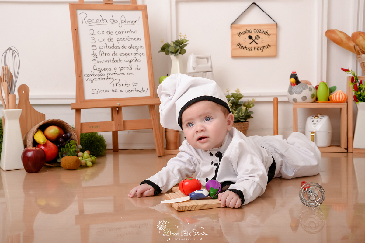 drica studio - ensaio fotográfico infantil - acompanhamento mensal- bebê vestindo roupa de chef de cozinha deitado de bruços no chão - cesto de frutas - cestos de pães - legumes de brinquedo - fundo branco com boiserie - piso amadeirado