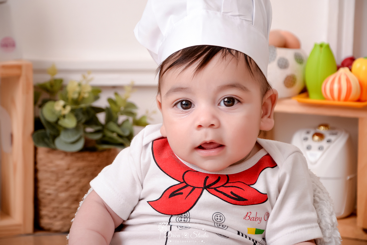 drica studio - ensaio fotográfico infantil - acompanhamento - bebê sentando em poltrona branca, vestindo camisa branca desenha um lenço - panela eletrica - legumes e galinha com ovos decorativo - cesto com planta - fundo branco