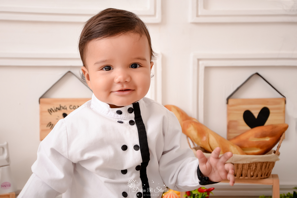 drica studio - ensaio fotográfico infantil - acompanhamento - bebê vestido roupa de chef de cozinha branca com botões e detalha preto - quadrinhos decorativo - cesto de pães - fundo branco com boiserie
