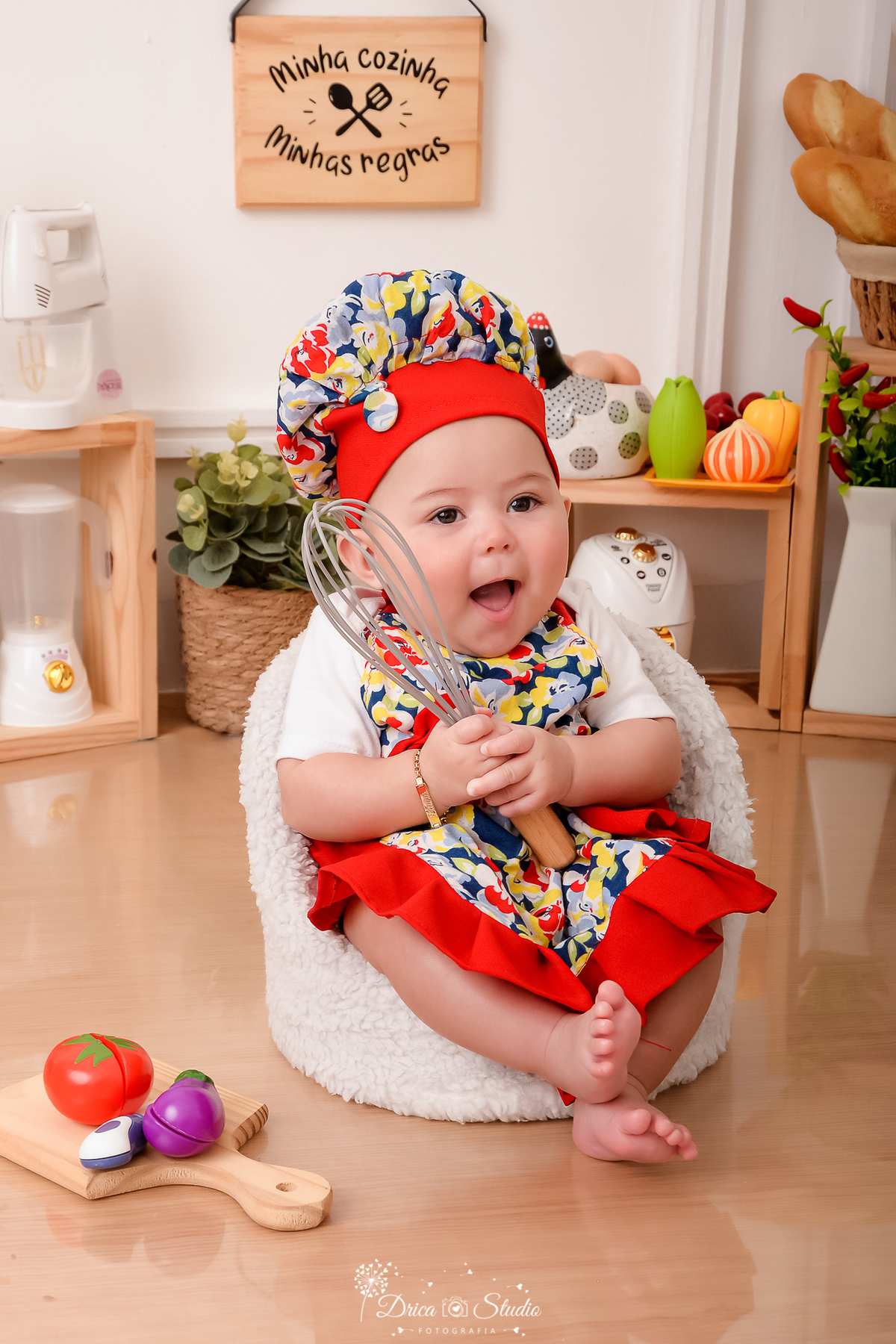 drica studio - ensaio fotográfico infantil - acompanhamento - bebê sentanda e poltrona, usando avental colorido com babado vermelho e chapéu de cozinheiro - segurado um batedor de massa - liquidificador, panela eletrica decorativa - cesto com plantas