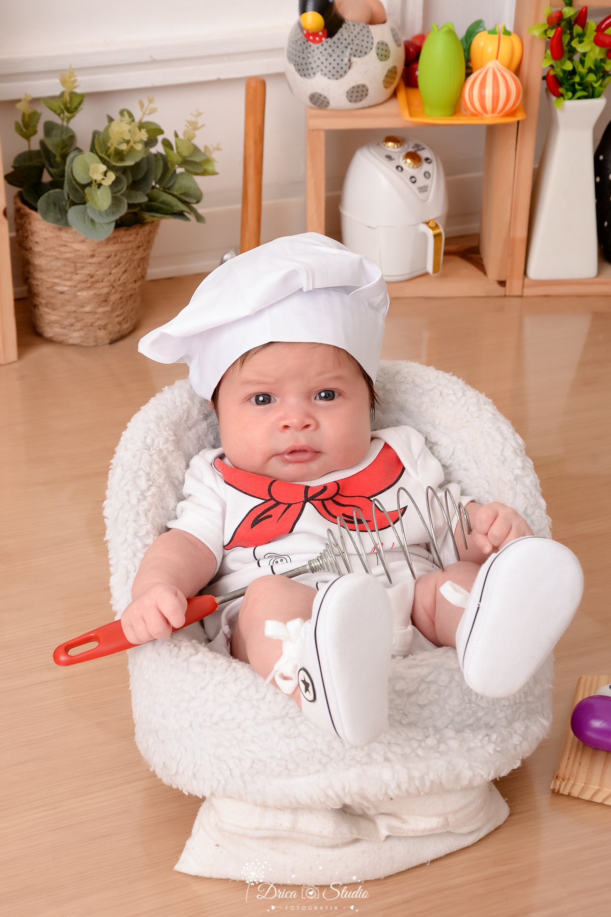 drica studio - ensaio fotográfico infantil - acompanhamento mensal- bebê sentando em poltrona branca - vestindo roupa de chef de cozinhal, segurando um batedor de massa - cestos de fruta - piso amadeirado - tenis branco