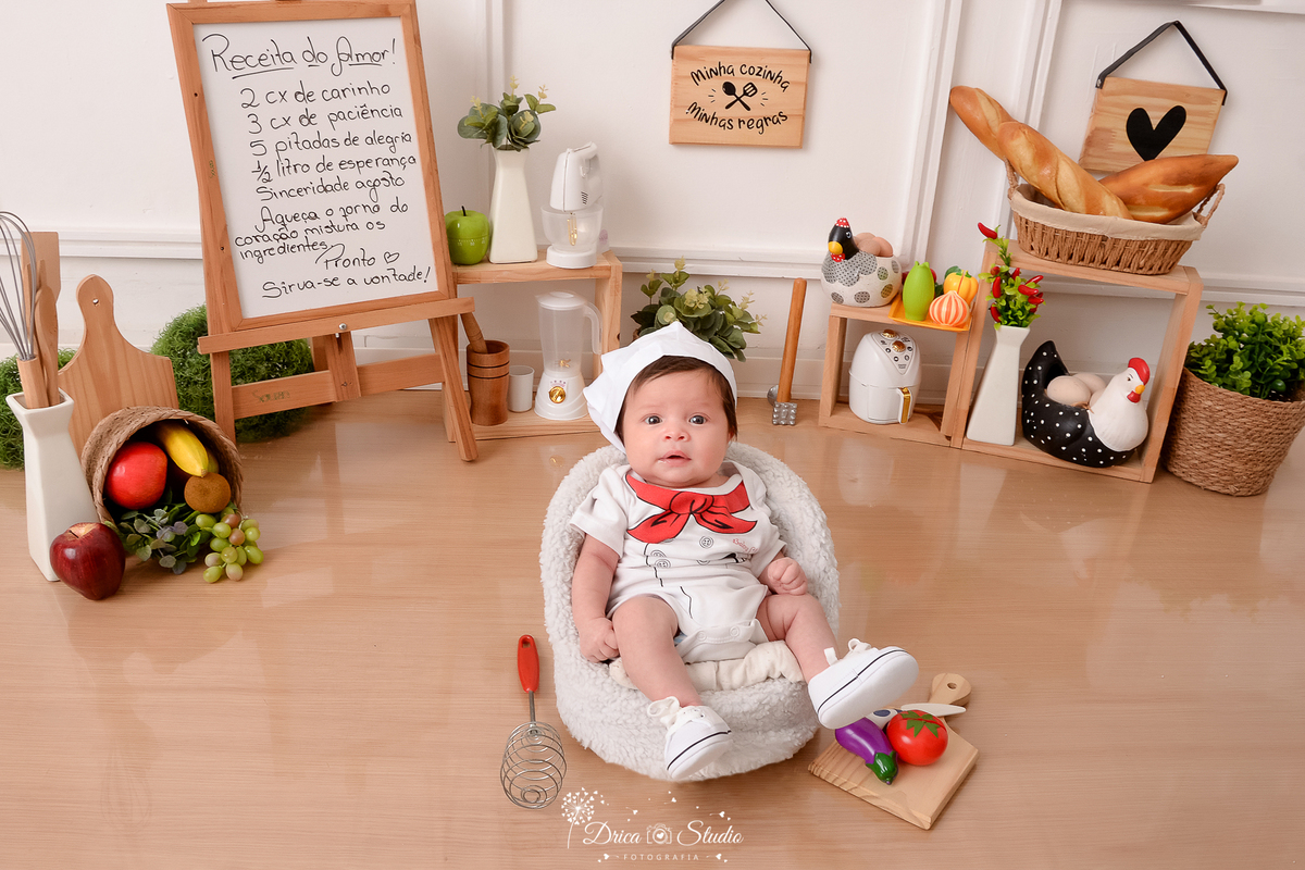drica studio - ensaio fotográfico infantil - acompanhamento mensal- bebê sentado em poltrona branca, vestindo como chef de cozinha, usando tênis branco - cesto com folhagem - cesto com pães - cestos com frutas - utensilios de cozinha - placas decorativas 