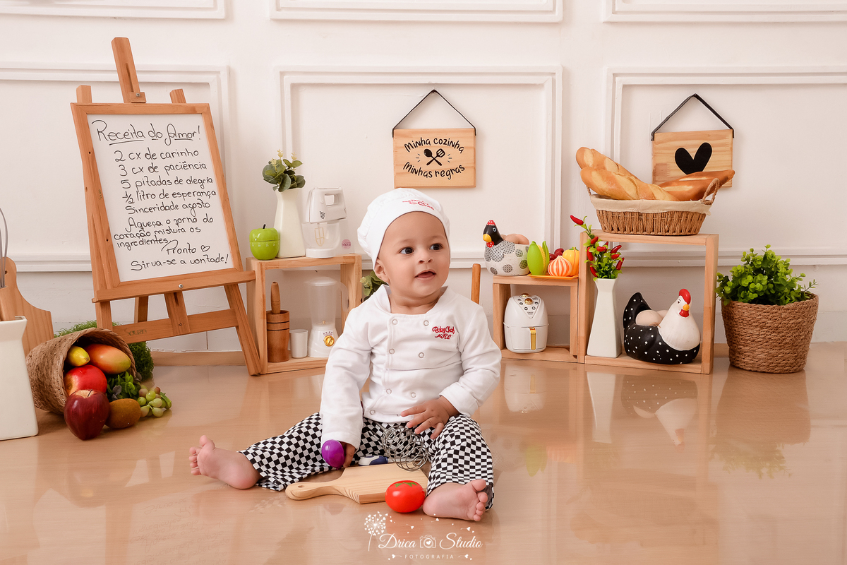 drica studio - ensaio fotográfico infantil - acompanhamento - bebê sentado no chão usando roupa de chef de cozinha - galinha, legume e pães decorativos - vaso com pimenteira - tabúa de cortar legumes - cesto com frutas - fundo branco com boiserie