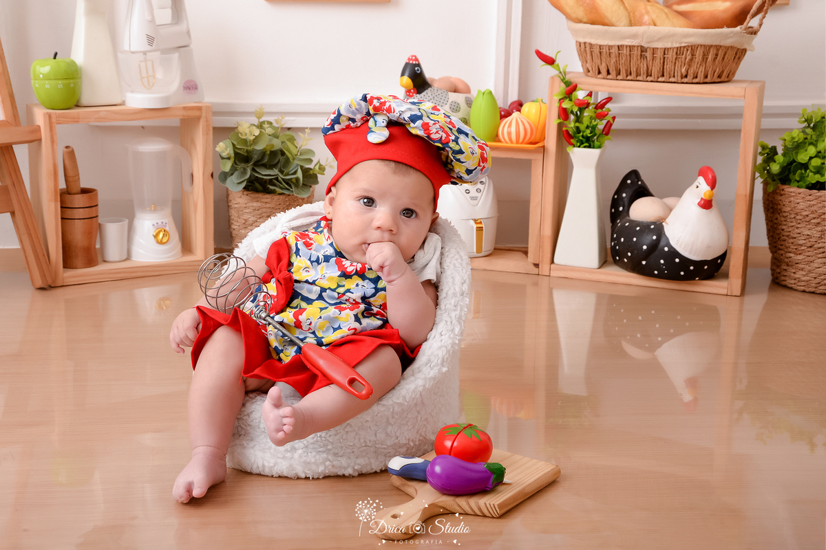 drica studio - ensaio fotográfico infantil - acompanhamento mensal- bebê sentada em poltrona, vestinda com roupas coloridas de chef de cozinha - galinha decorativa - utensilios de cozinha - vaso branco com folhagem - fundo branco com boiserie