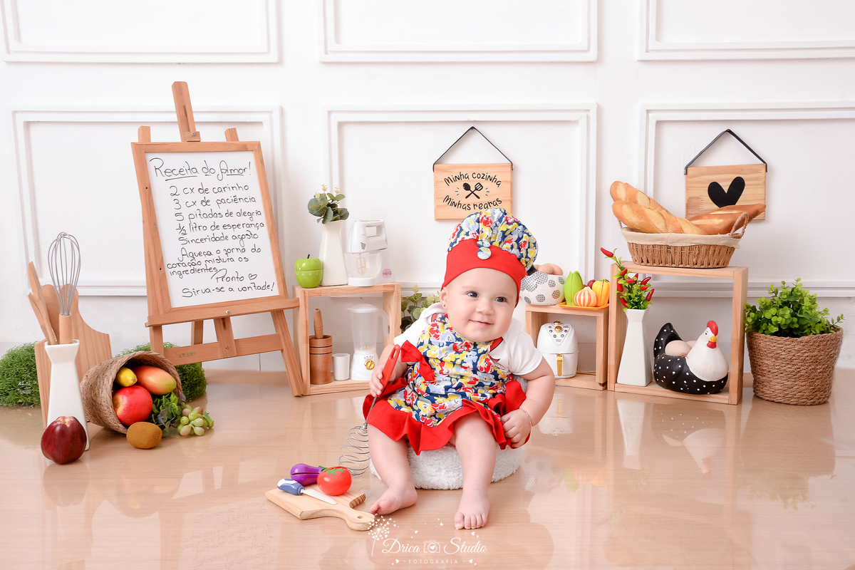 drica studio - ensaio fotográfico infantil - acompanhamento mensal- bebê sentada em poltrona, vestindo roupa de chef de cozinha estampada com detalhes vermelho - utensilios de cozinha, cesto com frutas, galinha decorativa - pães dentro de cesto 