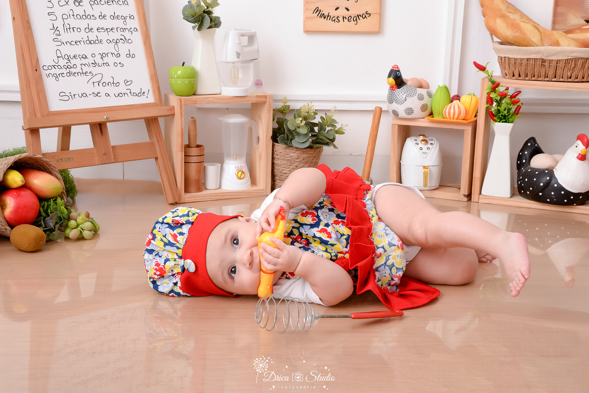 drica studio - ensaio fotográfico infantil - acompanhamento mensal- bebê usando avental e chapéu de chef de cozinha, deitada no chão, segurando um frango de borracha - cesto com frutas, cesto com pães - galinha decorativa - pilão- placa com receita 